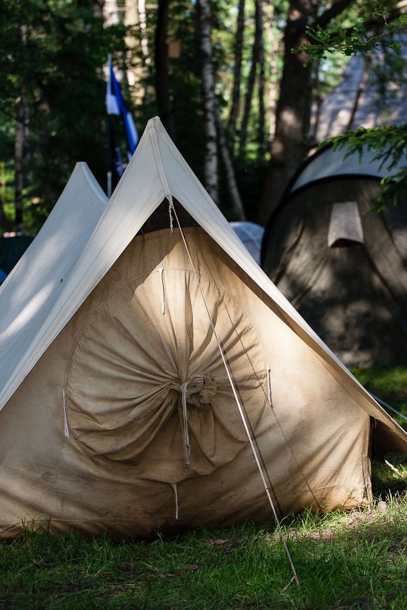 A beige canvas tent is set up on grassy terrain in a wooded area. The tent is closed at the front, with ropes securing the entrance. Another similar tent and trees are visible in the background, under sunlight filtering through the leaves. The scene suggests a campground reminiscent of Kilkim Žaibu 2012.
