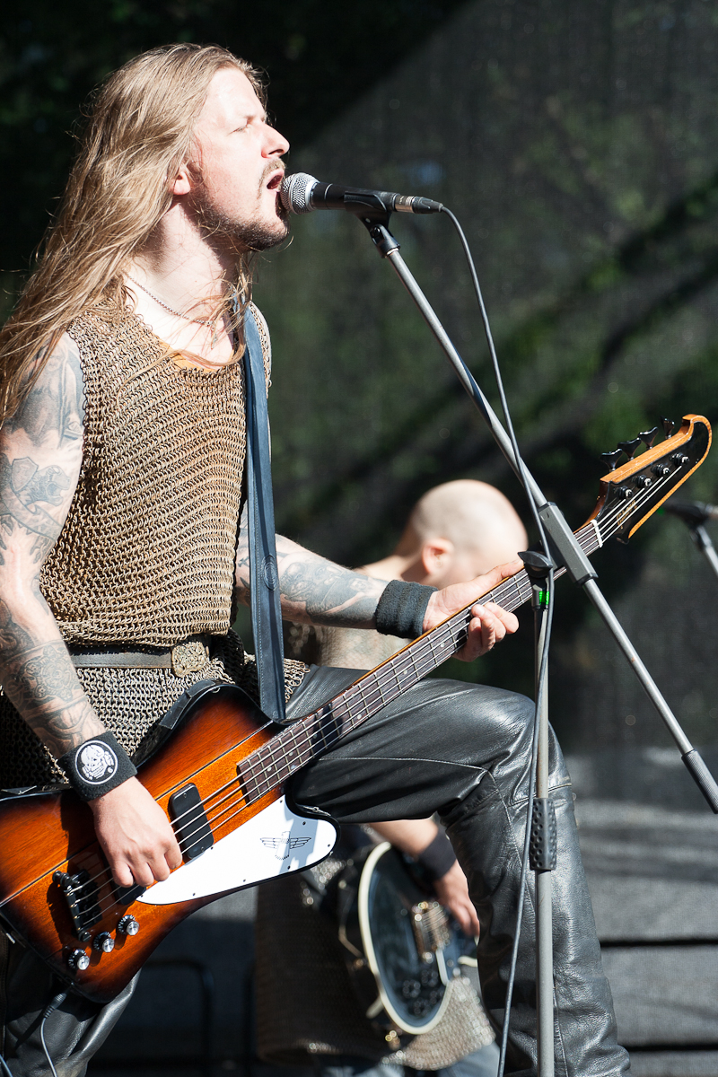 At the 2012 Kilkim Žaibu festival, a musician with long blonde hair and tattoos passionately sings into a microphone while playing a brown electric guitar on stage. He wears a chainmail shirt and black wristbands. Another band member with a shaved head is seen playing an instrument in the background.