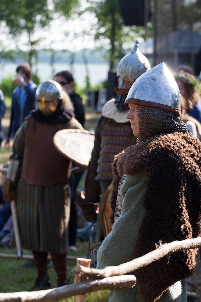 People dressed in medieval warrior costumes, featuring chainmail, helmets, and fur, stand outdoors among trees and grass. Some carry shields, while one focuses attentively to their right. The background is slightly blurred, showing other individuals and the natural setting of the 2012 Kilkim Žaibu festival.