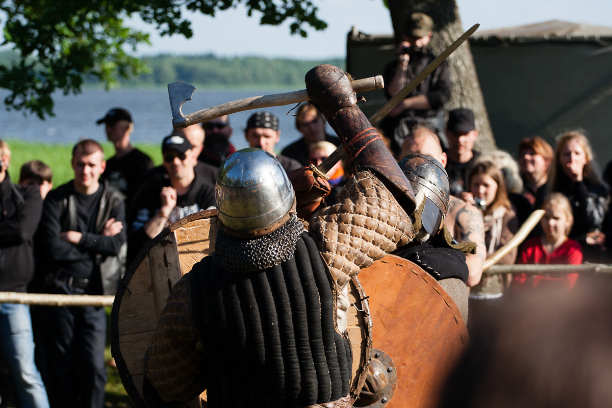 At a 2012 festival, two medieval re-enactors wearing chainmail and helmets engage in combat with shields and axes at an outdoor event. Onlookers in modern clothing surround them, watching intently. The backdrop includes a large tree, a tent, and the serene body of water visible in the distance.