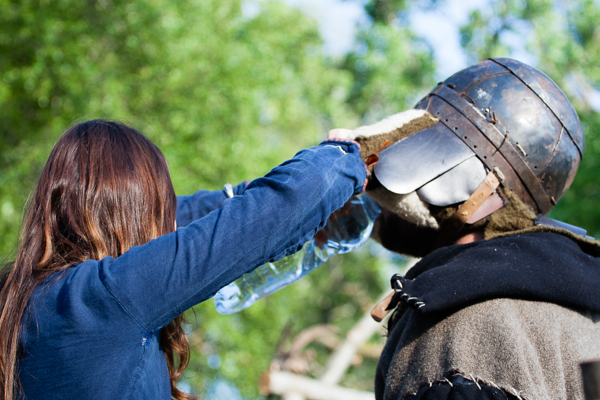 Attending the Kilkim Žaibu festival in 2012, a person with long brown hair, wearing a blue jacket, pours water from a plastic bottle over the mouthguard of a medieval helmet worn by another individual in armor. The background is blurred greenery, suggesting an outdoor setting.