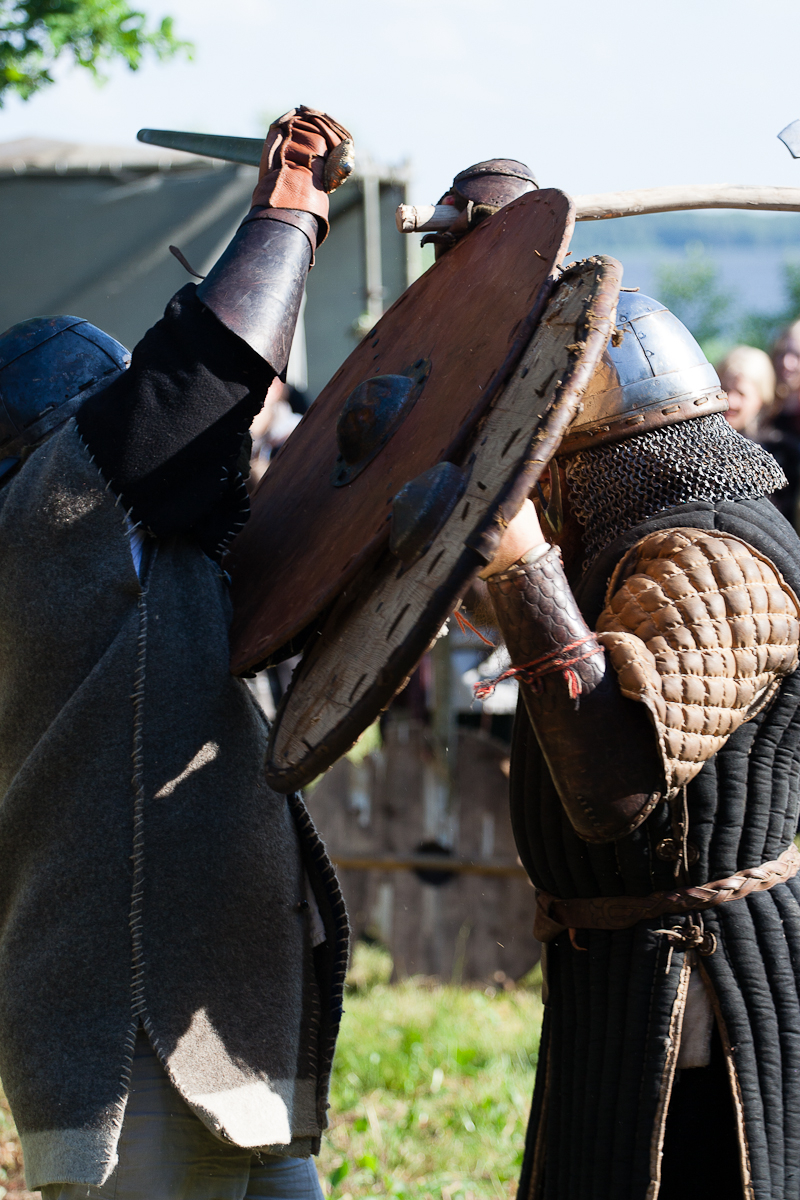 Two individuals engage in a medieval combat reenactment outdoors at Kilkim Žaibu 2012. Both wear period-appropriate clothing and metal helmets. They clash with one another; one raises a sword, and the other defends with a large round shield. Onlookers in the background watch the intense mock battle.