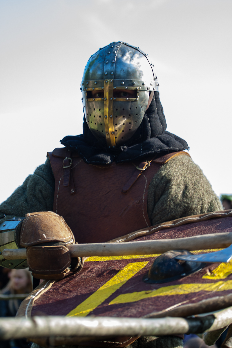 A person dressed in medieval armor, including a metal helmet with a visor, holds a shield adorned with yellow markings on a red background. Wearing a brown leather chest piece over a dark hood and green garment, the individual stands against a blurred outdoor background at the 2012 Kilkim Žaibu festival.