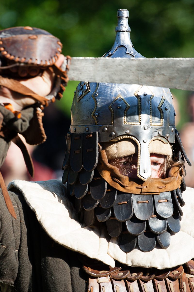 A person wearing medieval armor, including a scaled helmet with face protection and a padded collar, holds up a sword at Kilkim Žaibu 2012. The intricate helmet features metal plates and leather accents. The armor, detailed and well-crafted, gleams in the sunlight with a blurred outdoor background.