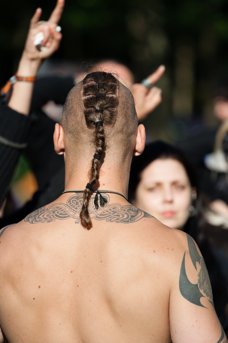 A shirtless person with intricate back tattoos and a long braided ponytail stands with their back to the camera at Kilkim Žaibu 2012. They are outdoors among a crowd, where some people are raising their hands. The background is slightly blurred, focusing on the person in the foreground and highlighting their hairstyle and tattoos.