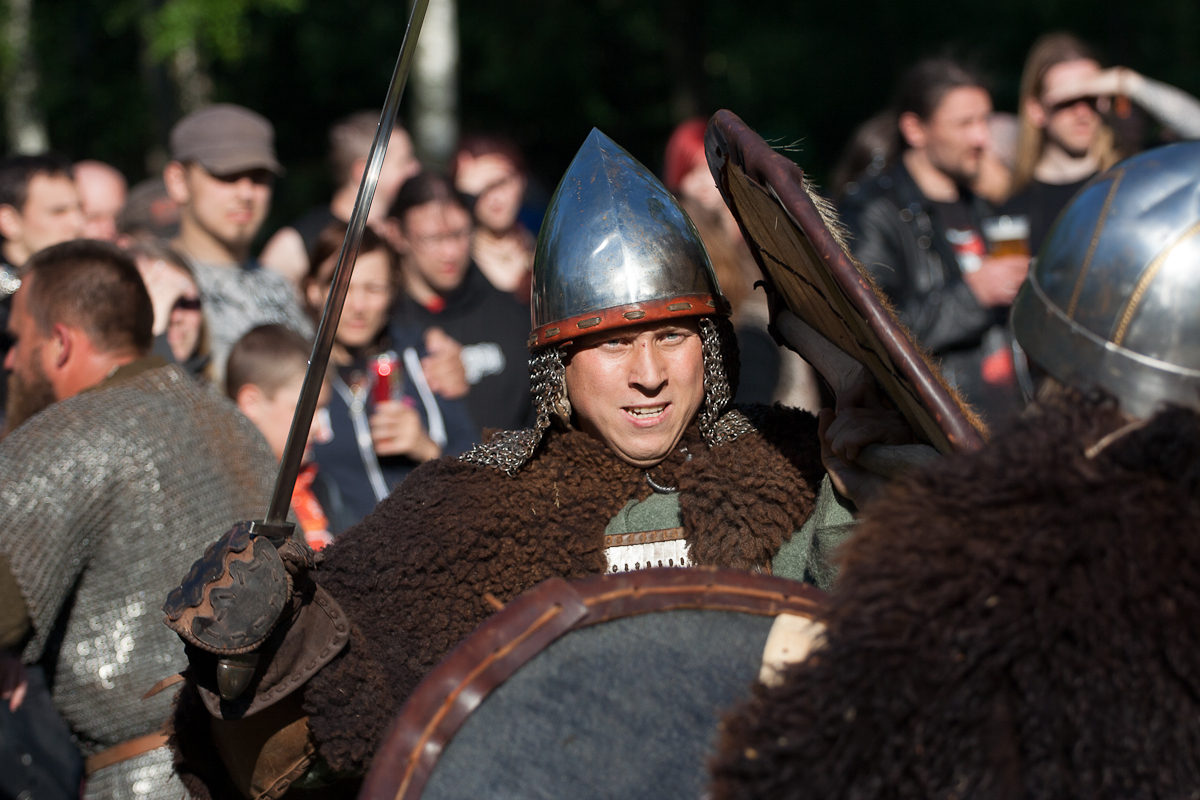 A person dressed as a medieval warrior, wearing a helmet and chainmail, holds a sword in one hand and a shield in the other. They appear to be in mid-battle during the 2012 Kilkim Žaibu reenactment event. The background shows a crowd of onlookers, many in casual clothing, observing the action.