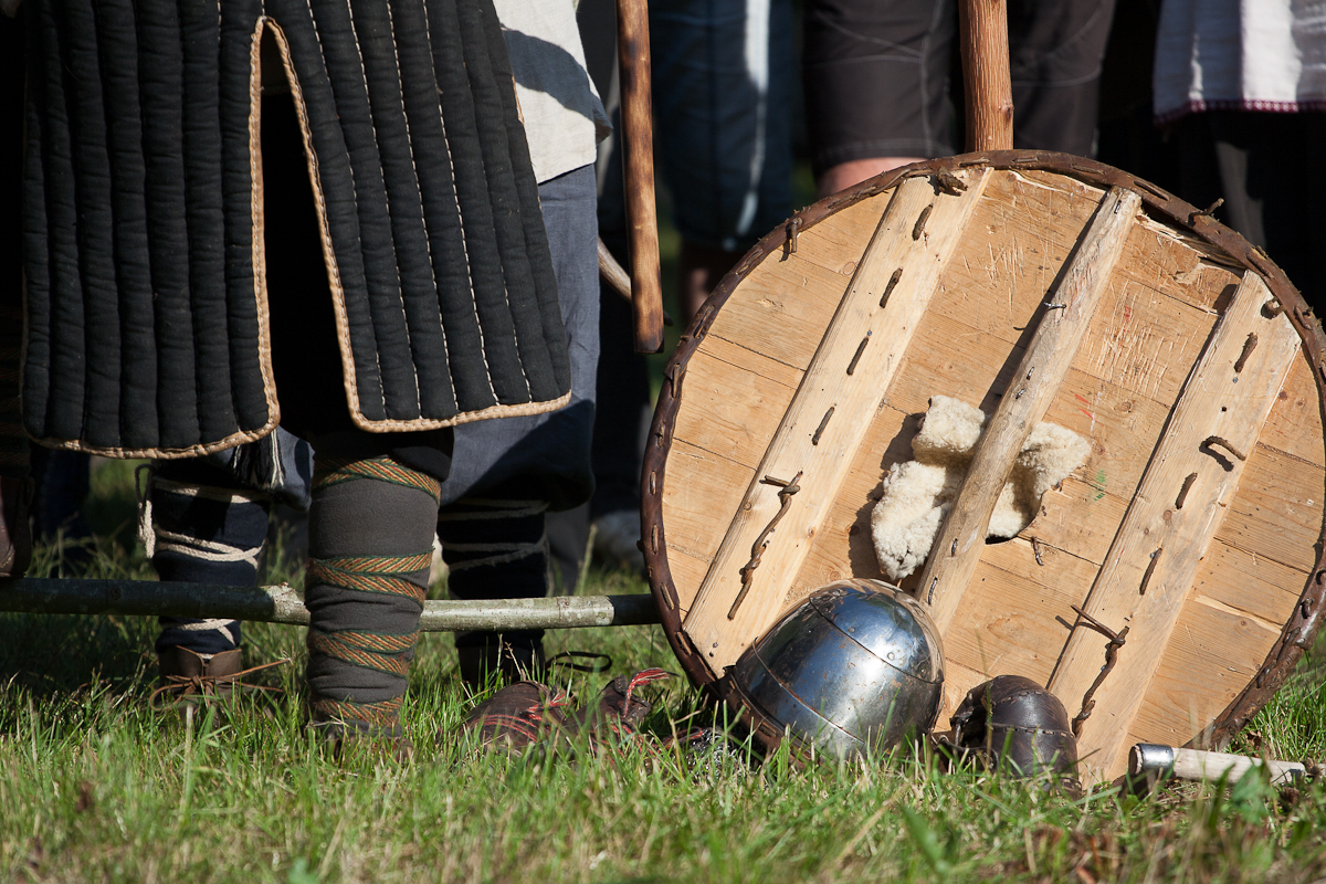 A medieval reenactor stands in a grassy field at Kilkim Žaibu 2012, with quilted leg armor and wrapped boots. At their feet are a wooden round shield, a metal helmet, and two small metal objects. The scene captures elements of historical combat attire and equipment.