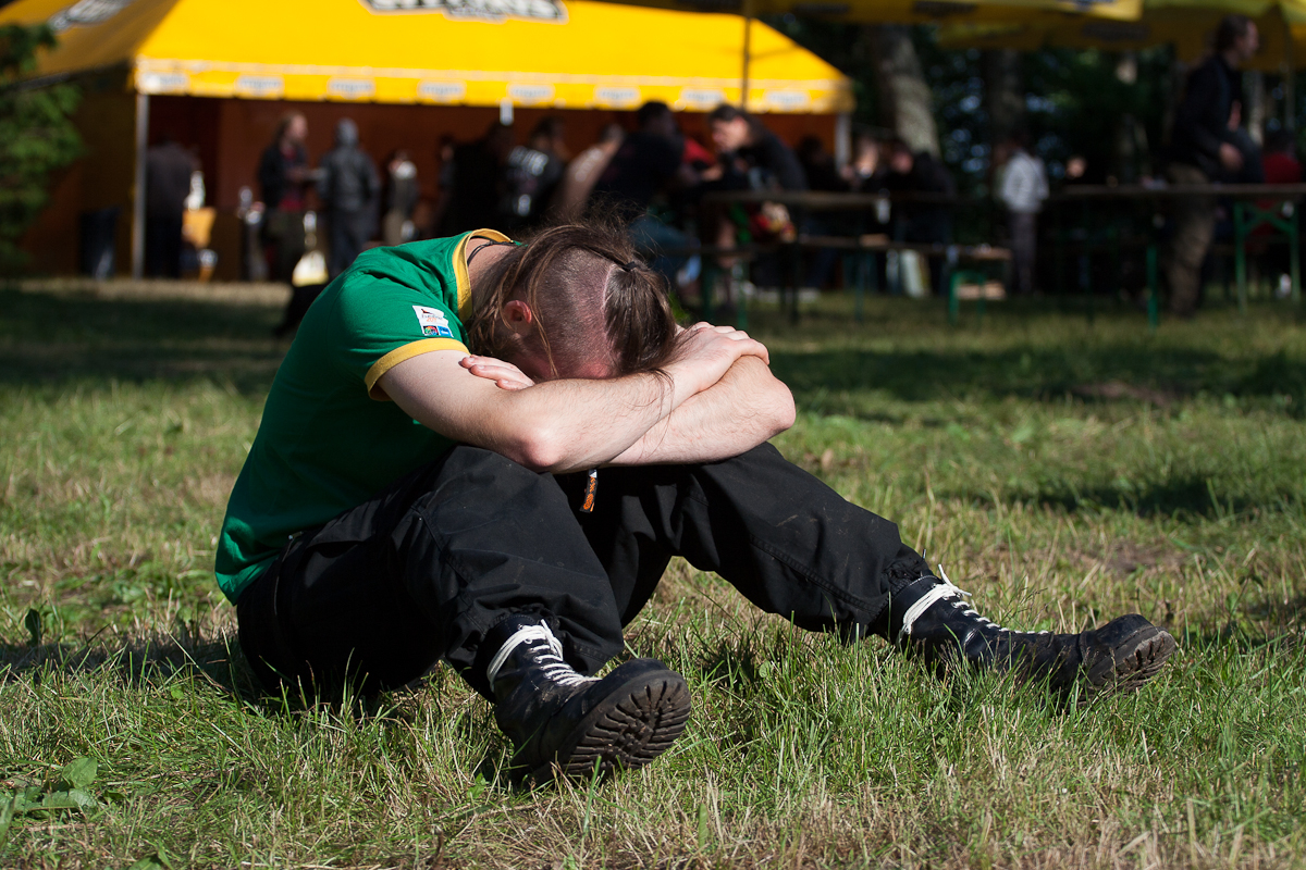 A person in a green shirt and black pants sits on the grass with their head resting on crossed arms and knees drawn up. The background features a yellow tent with "2012 Kilkim Žaibu" branded on it, and several people standing near it. The setting appears to be an outdoor event or festival.