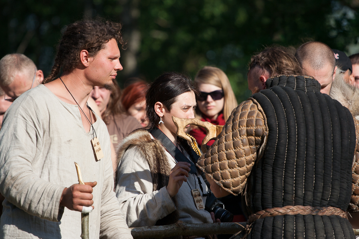 A group of people dressed in medieval clothing participate in the 2012 Kilkim Žaibu outdoor event. One man on the left holds a staff, while a woman in the center looks at a piece of parchment. Another person facing away from the camera wears a quilted garment with a rope belt. Other participants and onlookers are around.