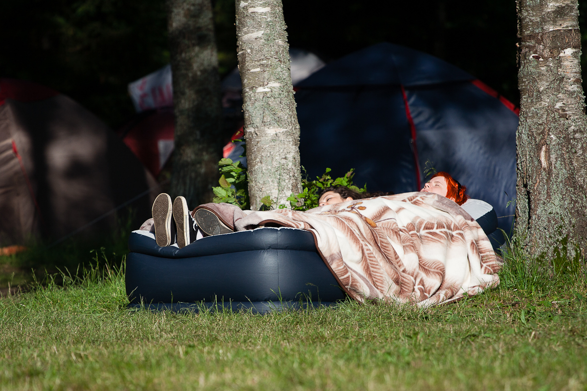 Two people are lying down on an inflatable mattress, covered with a blanket featuring leaf patterns. The mattress is placed outdoors on the grass, beneath trees with two tents visible behind them. The scene, reminiscent of Kilkim Žaibu 2012, captures the essence of a peaceful camping setting in daylight.