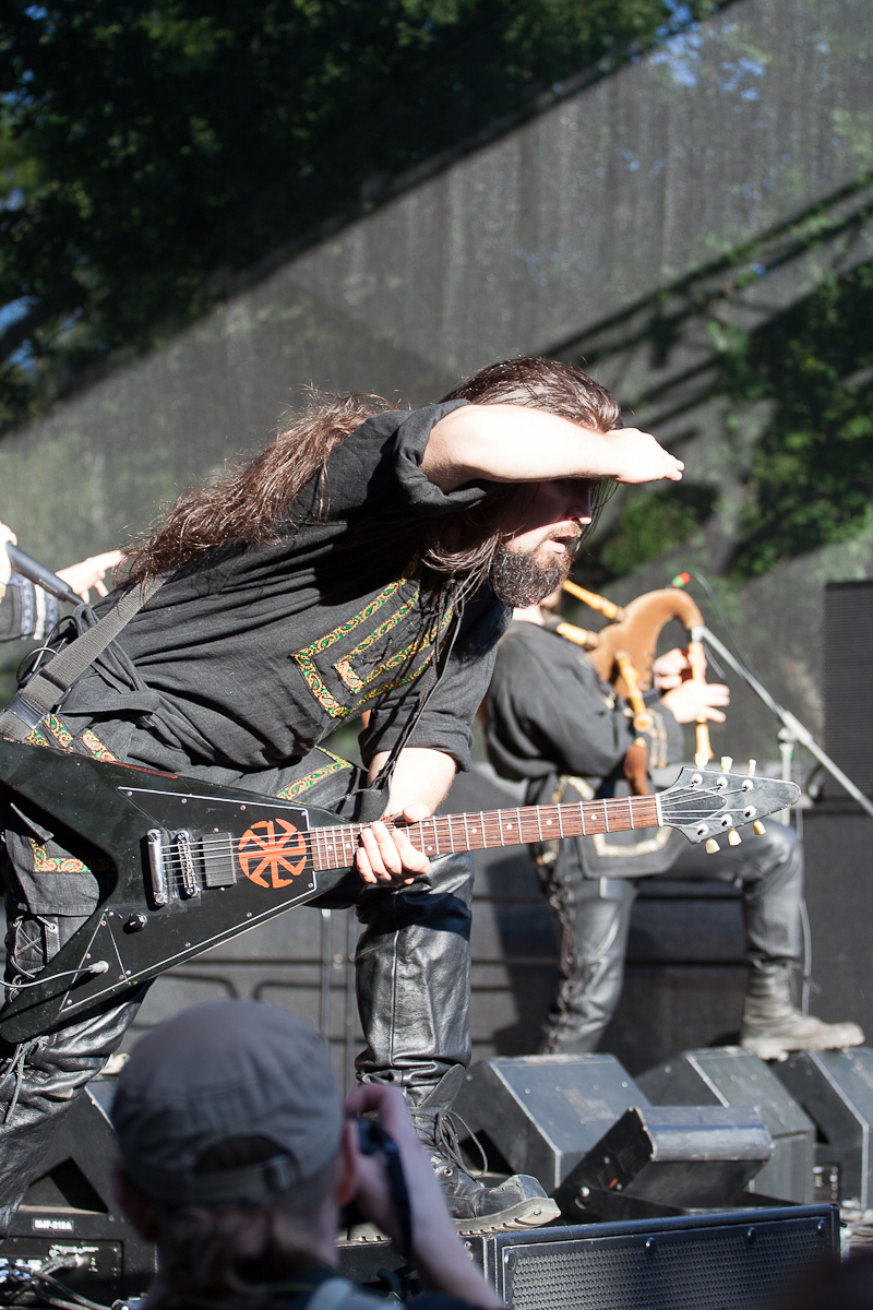 A musician with a long beard and long hair, wearing a black outfit, plays an electric guitar on an outdoor stage. He is shielding his eyes from the sun with his hand. Behind him, another musician plays the bagpipes. The audience at Kilkim Žaibu 2012 appears to be watching the performance intently.