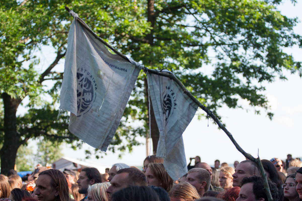 A crowd of people gathered outdoors beneath lush green trees at the 2012 Kilkim Žaibu festival. Two white flags with blue circular symbols are attached to a single branch held above the crowd. The sky is clear, creating a bright and cheerful atmosphere at the gathering.