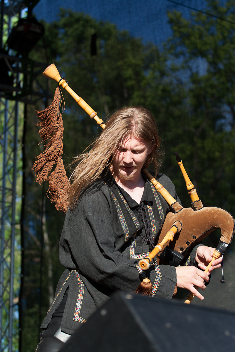 A musician with long blond hair plays the bagpipes on an outdoor stage at Kilkim Žaibu 2012. They wear a black, traditionally styled tunic with decorative patterns. The bagpipes feature a wooden, natural finish and a decorative tassel. Trees and stage lighting are visible in the background.