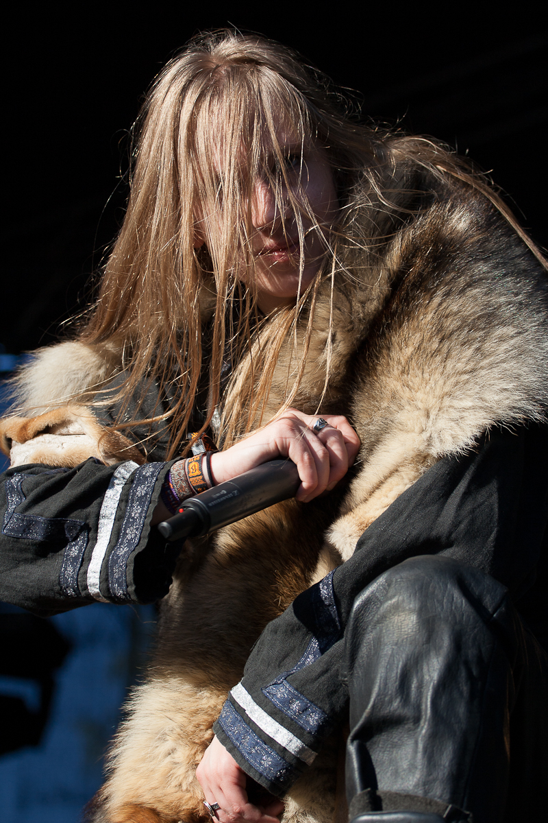 A person with long hair obscuring part of their face is holding a microphone close to their mouth. They are wearing a fur garment over a dark, intricately decorated outfit. The background is dark, with a slight glimpse of blue sky, reminiscent of the night at the 2012 Kilkim Žaibu festival. The intense expression suggests they are performing passionately.