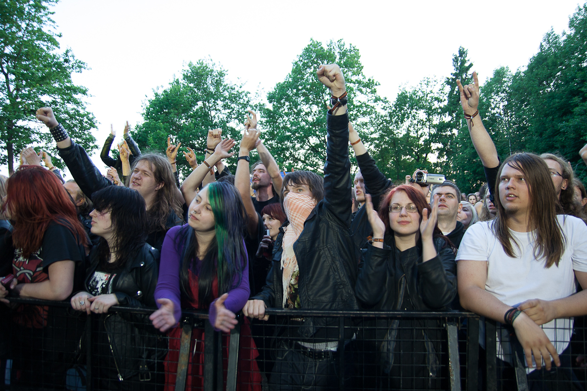 A large crowd of enthusiastic concertgoers stands behind a barricade at the 2012 Kilkim Žaibu festival. People are dressed in casual clothing and leather jackets, some with dyed hair and accessories. Many raise their fists in the air, cheering energetically. Trees and a clear sky are visible in the background.
