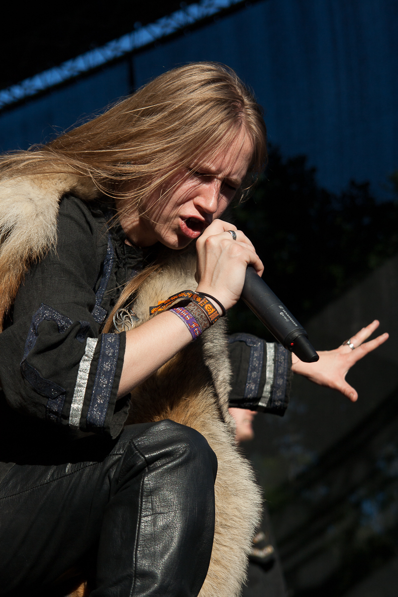 A singer with long blond hair performs passionately on stage at Kilkim Žaibu 2012, holding a microphone close to their mouth. They wear a dark top with patterned trim, leather pants, and several colorful wristbands. A fur stole drapes over their shoulders, and a dark backdrop with foliage is visible behind them.