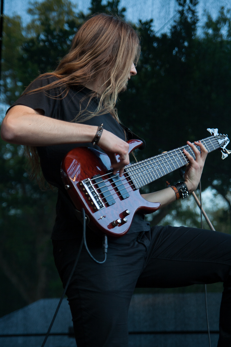 A person with long, light brown hair plays a six-string electric bass guitar at the 2012 Kilkim Žaibu festival. They are wearing a black shirt and black pants, and wristbands on both wrists. The background is an outdoor setting with trees blurred in the distance. The person seems intensely focused on the instrument.
