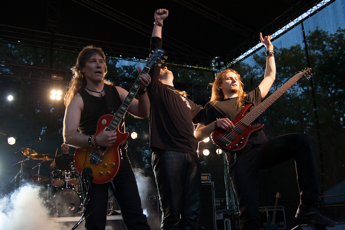 At the 2012 Kilkim Žaibu festival, three male musicians perform energetically on an outdoor stage. The one on the left plays an electric guitar, the one on the right a bass guitar, and the middle musician raises his fist. A drummer is seen in the background. Stage lights shine, and fog creates a dramatic atmosphere.