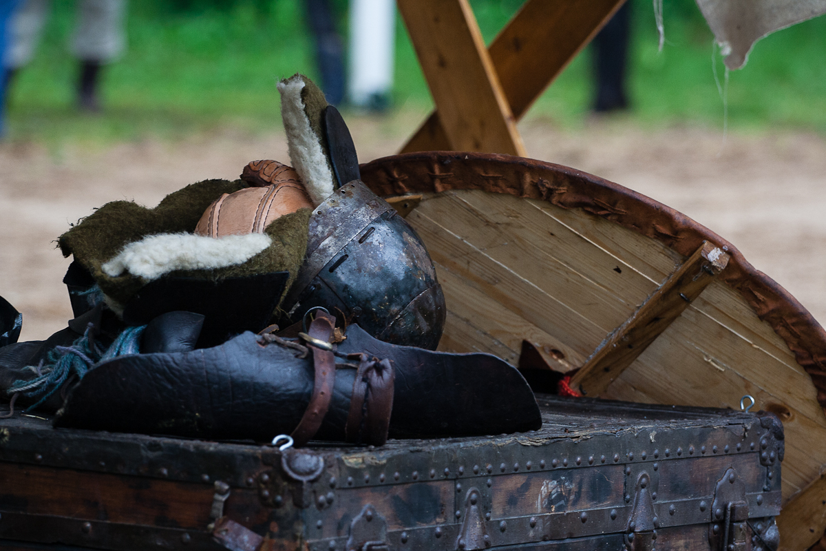 A medieval helmet, fur-lined leather armor pieces, and a wooden shield rest on a weathered trunk at the 2012 Kilkim Žaibu festival. The scene is set outdoors with grass and a blurred background, suggesting a historical reenactment or period setting. Wooden beams behind the trunk form a partial structure.