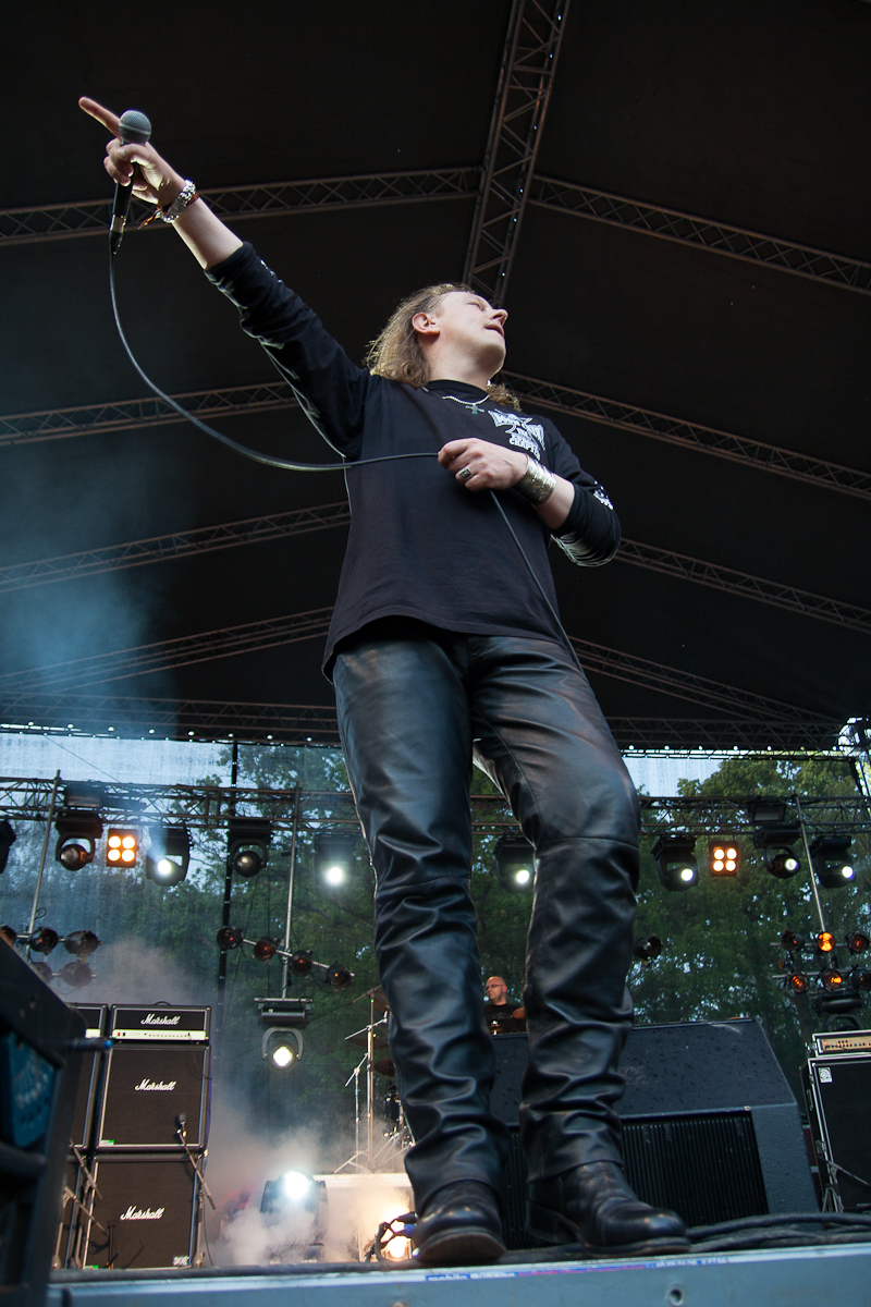 Lead singer holding a microphone and performing energetically on an outdoor stage at the 2012 Kilkim Žaibu festival. The singer is wearing a black long-sleeve shirt and black leather pants. Stage lights and amplifiers can be seen in the background, with trees visible beyond the stage structure. The scene captures a dynamic moment of the performance.