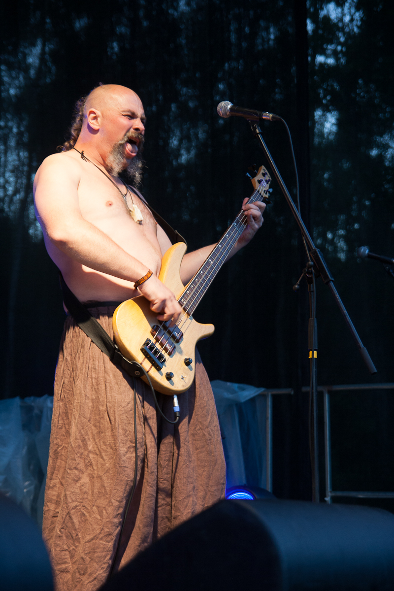 A shirtless man with a bald head and long braided ponytail passionately plays a bass guitar on stage at Kilkim Žaibu 2012. He is wearing brown, loose-fitting pants and has several accessories on his wrists. Singing or shouting into a microphone, the dark background is lit by a faint blue glow.
