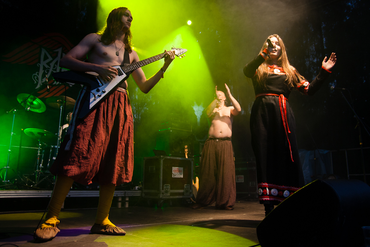 On a dimly lit stage at the 2012 Kilkim Žaibu festival, a barefoot guitarist in a long skirt plays next to a singer in a black and red traditional-style dress holding a microphone. In the background, a shirtless man in a long skirt passionately claps his hands. A drum set is visible on the left side of the stage.
