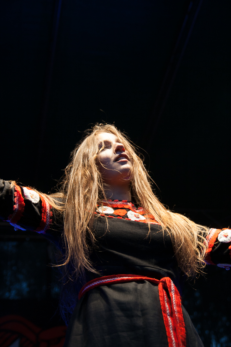 A person with long, flowing hair stands on a dimly lit stage at the 2012 Kilkim Žaibu festival, with arms outstretched. They are wearing a black garment adorned with red and white floral patterns and a red belt. The lighting casts dramatic shadows, emphasizing their expressive pose against the dark background.
