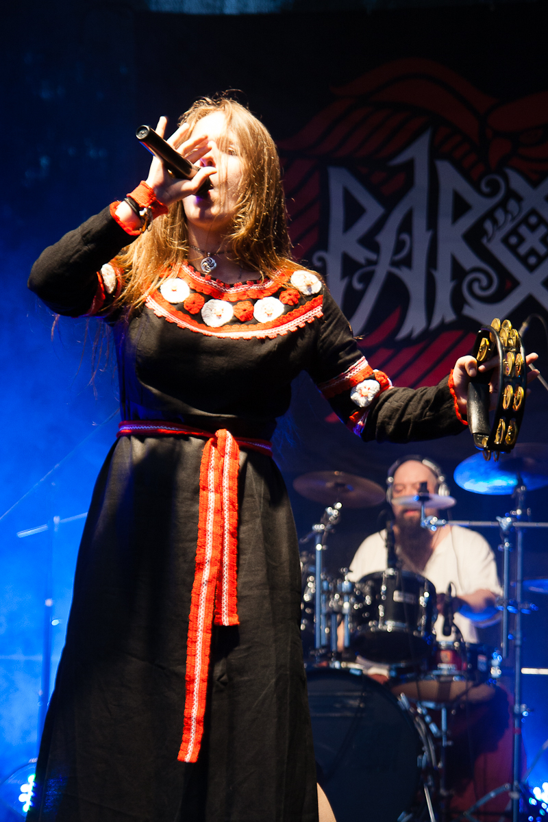 A woman in a black dress with red and white embroidery sings passionately into a microphone on stage at Kilkim Žaibu 2012. Her long hair flows over her shoulders as she holds a tambourine in her left hand. A drummer performing in the background is partially visible, with stage lights creating a blue illumination around them.