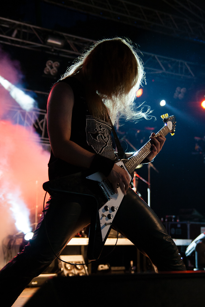 A guitarist with long hair plays an electric guitar energetically on a dimly lit stage at Kilkim Žaibu 2012. He is wearing a sleeveless black shirt and black leather pants. Smoke and colorful stage lights create a dramatic atmosphere. The guitar has a white body, and his hair obscures his face as he leans forward.