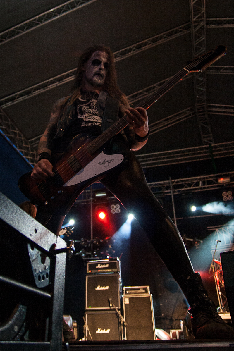 A rock musician with a painted face and long hair plays an electric bass guitar onstage at the 2012 Kilkim Žaibu festival. He wears a sleeveless shirt and tight black pants, standing with legs apart under a large stage with dramatic lighting and visible rigs and amplifiers behind him.
