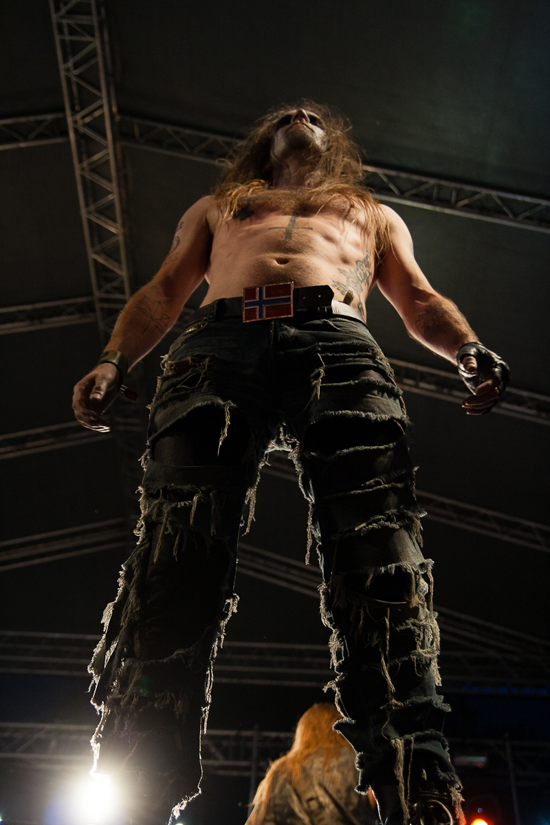 A muscular, shirtless man with long hair stands under stage lighting, seen from a low angle. He wears tattered black pants, black wristbands, and a belt adorned with a flag resembling Norway's. Amidst the backdrop of metal framework and stage equipment at Kilkim Žaibu 2012, he appears deep in performance.