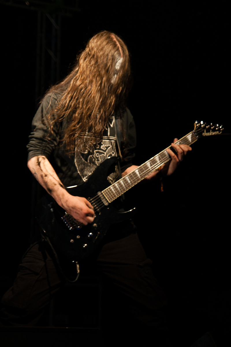 A musician with long, flowing hair obscuring their face is playing an electric guitar with intensity at the Kilkim Žaibu festival. Dressed in dark clothing and deeply focused on their performance on stage against a dark background, they wield a black and white designed guitar, capturing the essence of 2012.