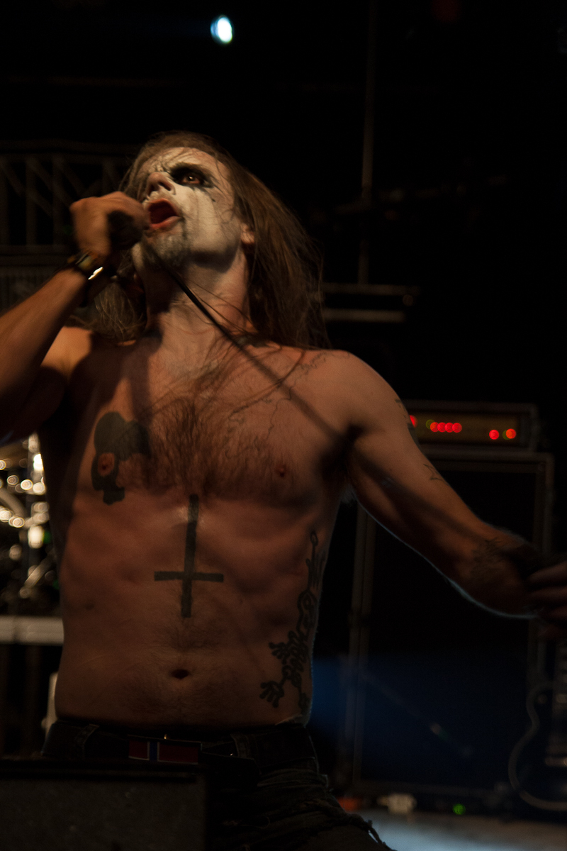 At the Kilkim Žaibu festival in 2012, a shirtless man with long hair performs on stage, holding a microphone to his mouth with one hand. His face is painted white with dark makeup around his eyes and mouth. He has tattoos, including an inverted cross on his torso. The background is dark and filled with stage equipment.