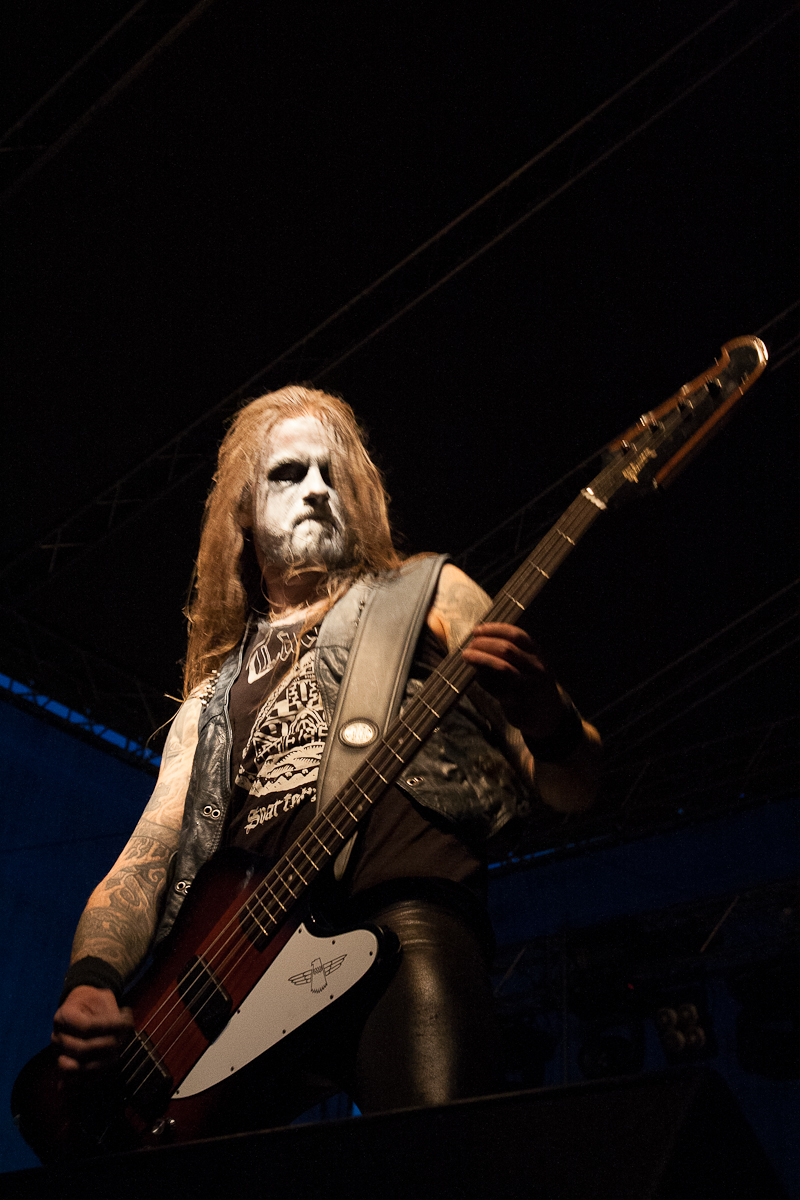 A musician with long hair and face paint plays a bass guitar on a dimly lit stage at Kilkim Žaibu festival in 2012. He wears a sleeveless denim vest over a black shirt with intricate designs. The background is dark, with subtle stage lighting hinting at a live performance setup.