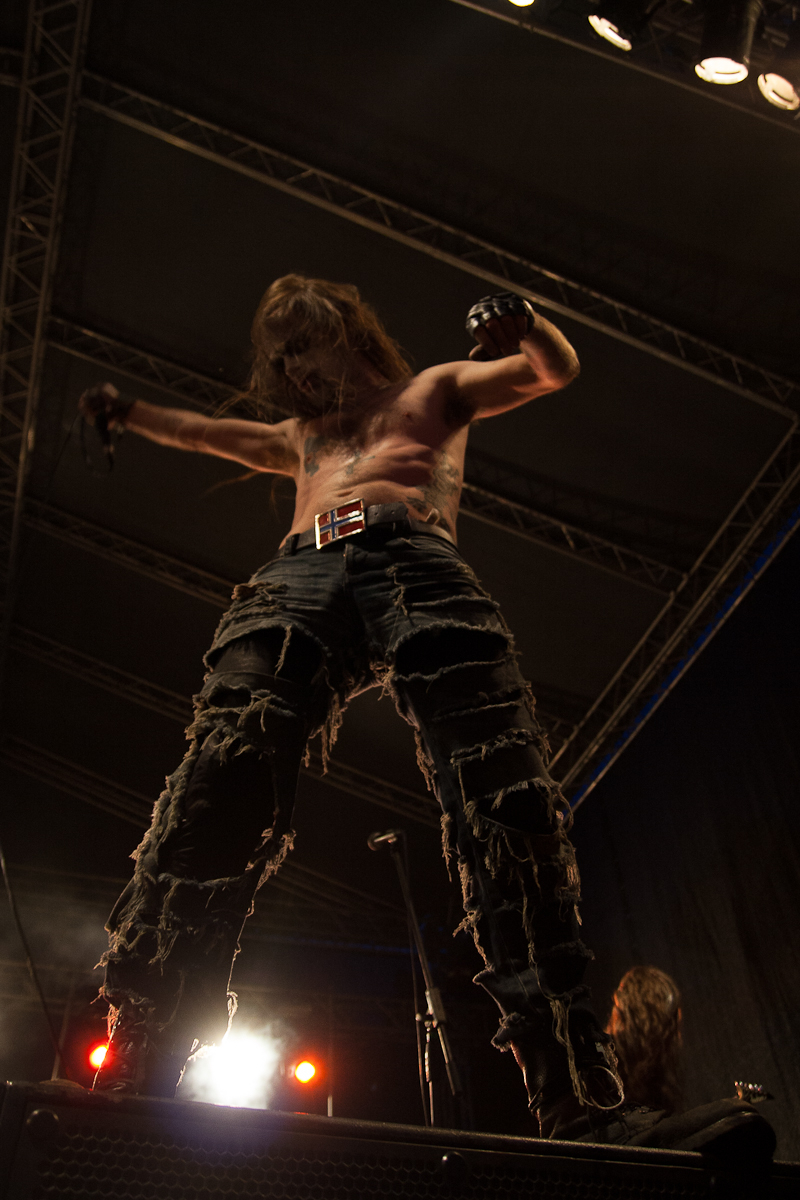 At the 2012 Kilkim Žaibu festival, a shirtless performer on stage stands with arms raised, holding a microphone. He wears distressed, ripped jeans with a belt and fingerless gloves. His long hair obscures his face. Stage lights illuminate him from below, highlighting the dramatic pose and creating an intense atmosphere.