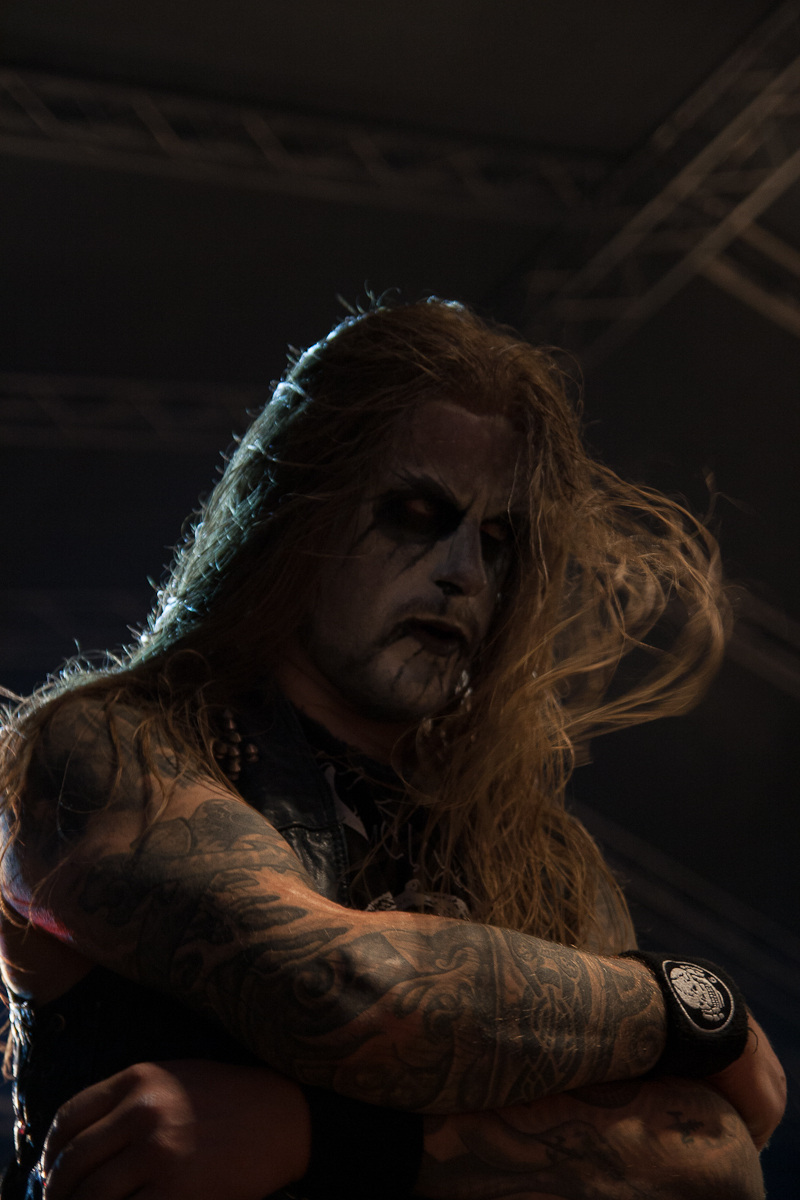 A man with long, flowing hair and heavy black and white face paint stands under stage lighting at the 2012 Kilkim Žaibu festival, arms crossed. He wears a sleeveless outfit, showcasing his tattoo-covered arms, and has a serious expression with a distant gaze. The background is dark, with indistinct lighting structures.
