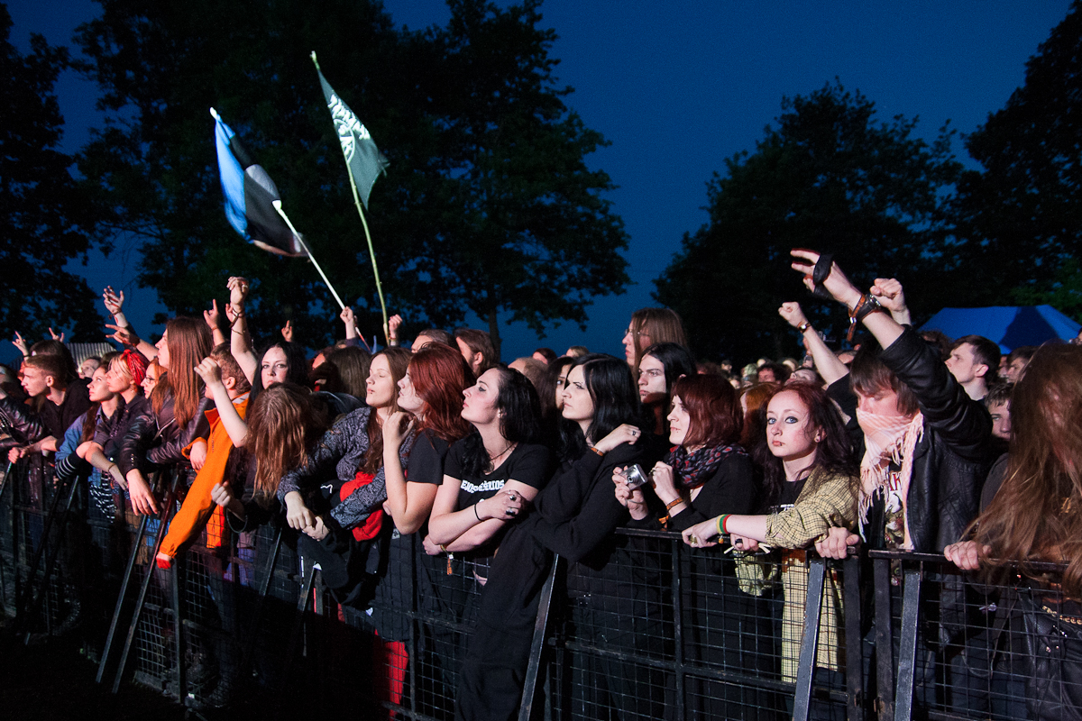 A crowd of excited people at an outdoor concert at dusk, many with raised arms, some holding flags in the background. They are standing behind a metal barrier with trees silhouetted against a darkening sky behind them. The atmosphere is lively and energetic, reminiscent of Kilkim Žaibu 2012.