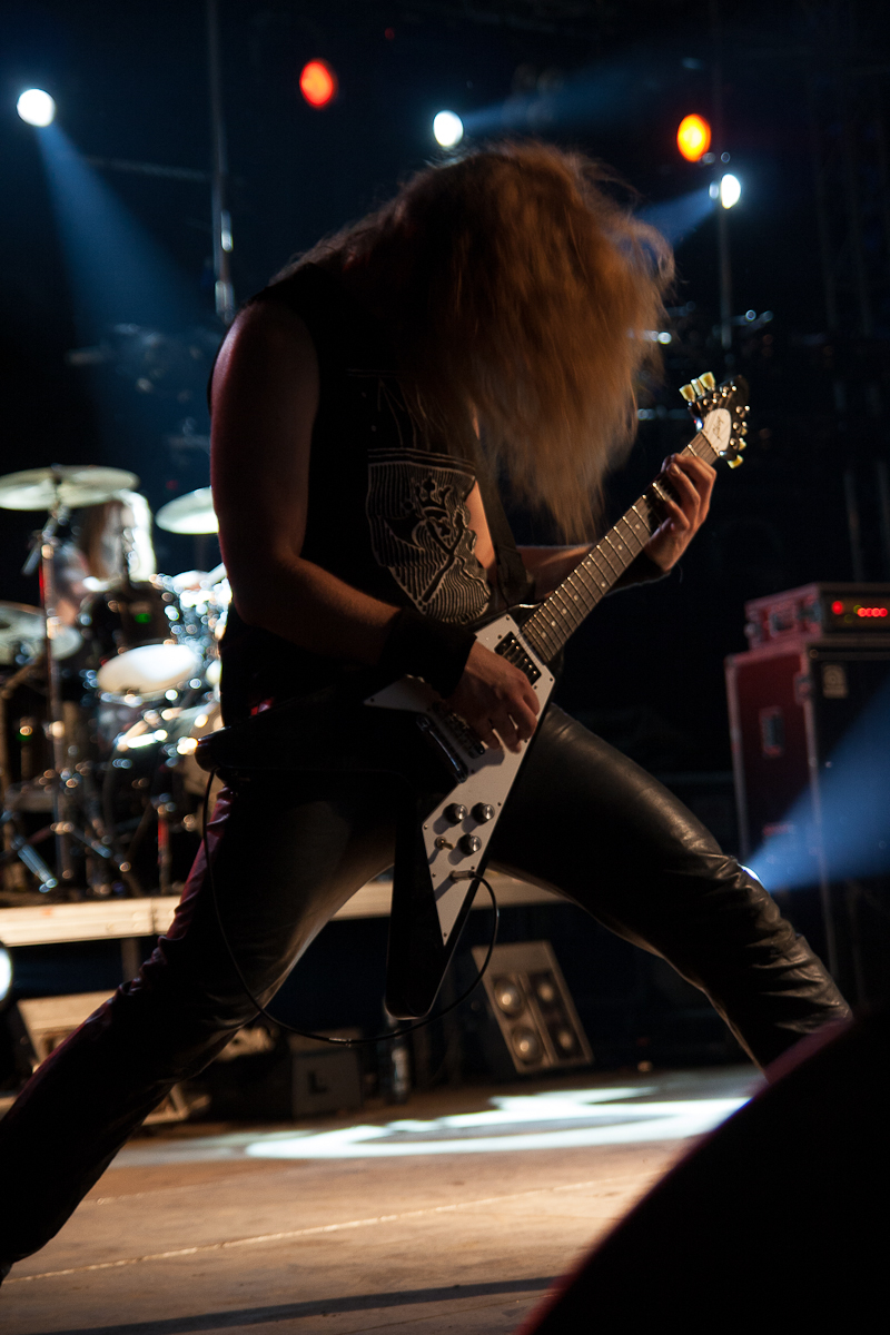 A musician with long hair in motion plays an electric guitar on stage at the 2012 Kilkim Žaibu festival, wearing a sleeveless shirt and black leather pants. In the background, a drummer plays a drum set. The stage is illuminated by vibrant blue and white lights, creating a dynamic atmosphere.