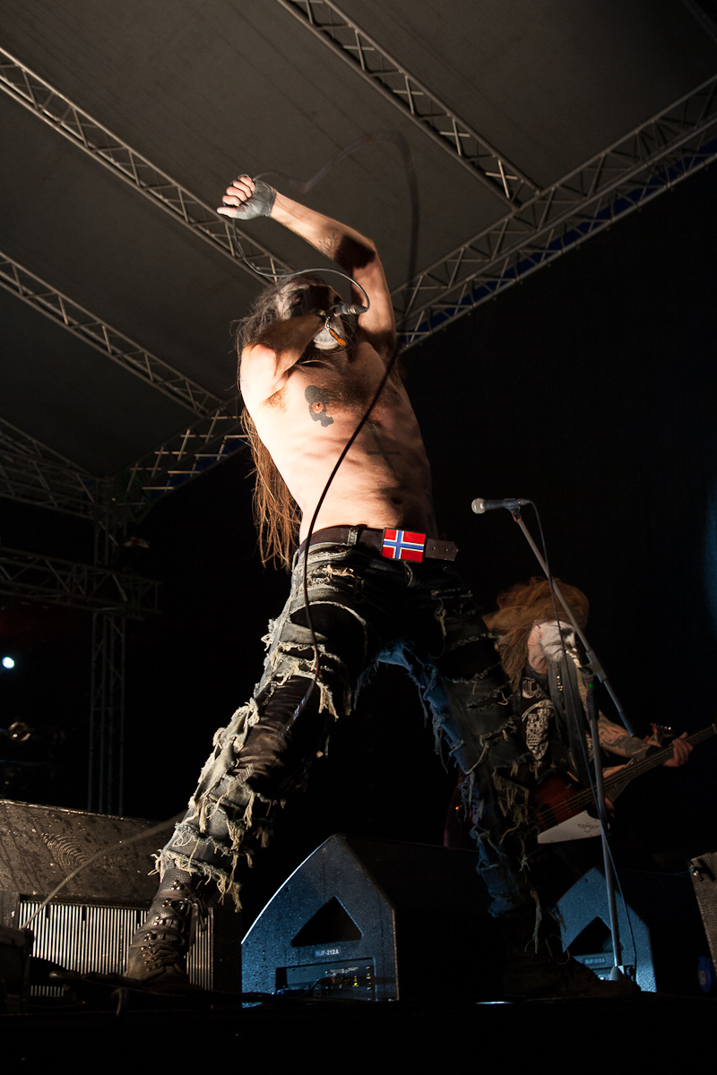 A shirtless musician with long hair performs on stage at Kilkim Žaibu 2012, wearing torn black jeans and a belt with a Norwegian flag buckle. He has white face paint with black details and energetically raises his arms, holding a microphone. Another band member plays guitar in the background under stage lights.