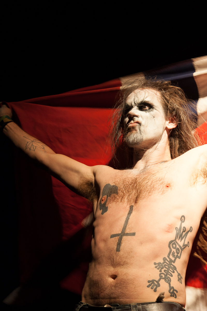 A shirtless person with long hair and face paint stands with arms raised, holding a red and black flag behind them. They have tattoos, including a large cross on their torso and various others on their arms and chest. The dark background highlights their dramatic appearance, resembling the fervor of the Kilkim Žaibu festival.
