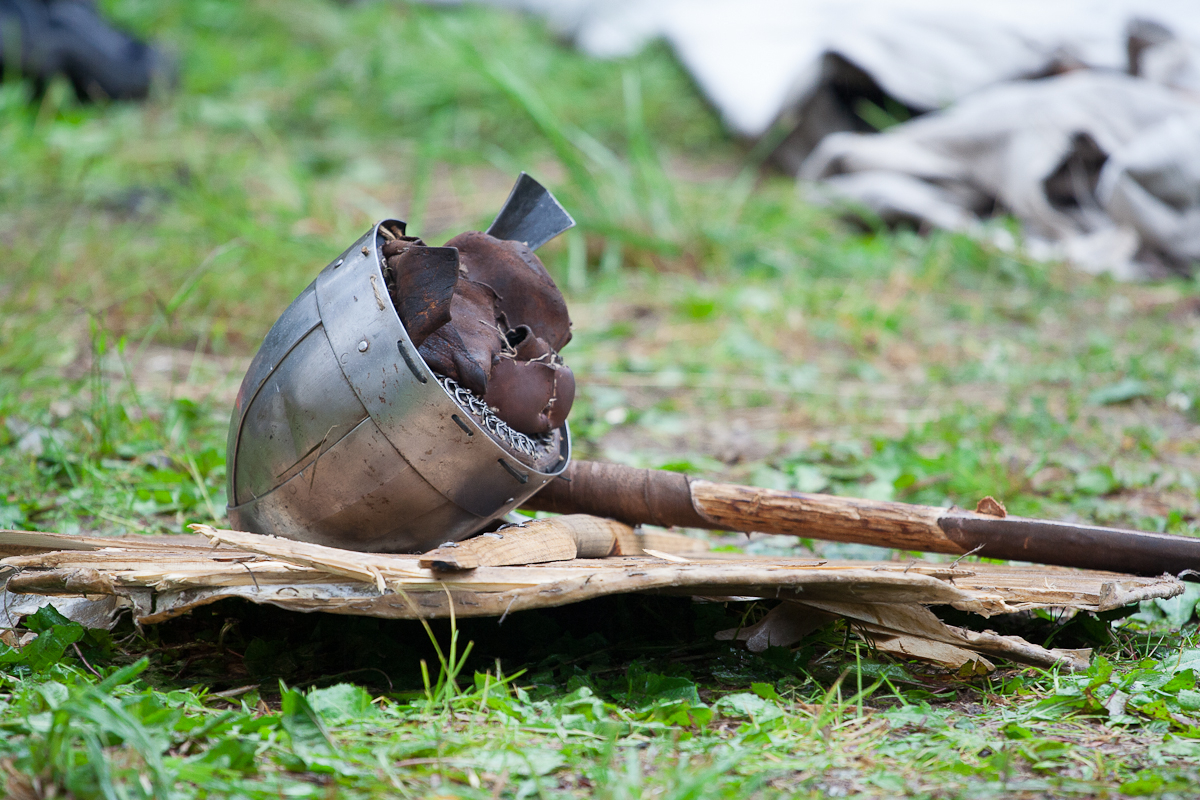 A medieval-style helmet and a wooden-handled weapon, possibly a mace or axe, rest on a makeshift wooden surface outdoors with greenery in the background. The scene suggests a historical reenactment or display of medieval artifacts at Kilkim Žaibu 2012, with a soft focus on the background elements.