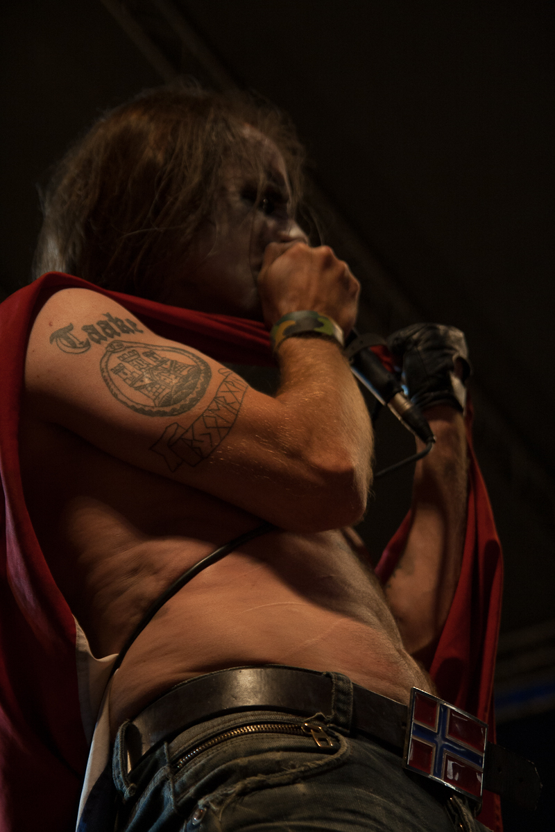 A singer with long hair and face paint holds a microphone close to his mouth. He is shirtless, revealing a tattoo on his right bicep. Wearing a red cape and a belt with a buckle displaying the Norwegian flag, he performs at the 2012 Kilkim Žaibu festival. The dark background suggests an indoor or nighttime performance.