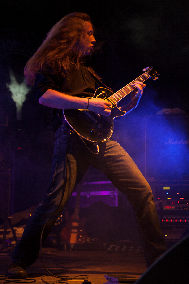 At Kilkim Žaibu 2012, a guitarist with long hair performs energetically on stage, illuminated by dramatic lighting. He plays a black electric guitar and is dressed in a black shirt and blue jeans. Amplifiers, including one from Marshall, are visible in the background, surrounded by blue and purple stage smoke.
