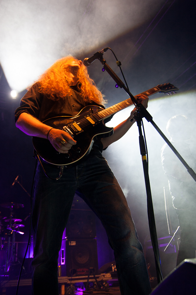 A musician with long, wavy blond hair and a beard performs on stage with a black electric guitar. Wearing a black shirt and blue jeans, he stands under dramatic stage lighting, casting a smoky, atmospheric glow. A microphone is positioned in front of him amid band equipment—evoking the raw energy of Kilkim Žaibu 2012.