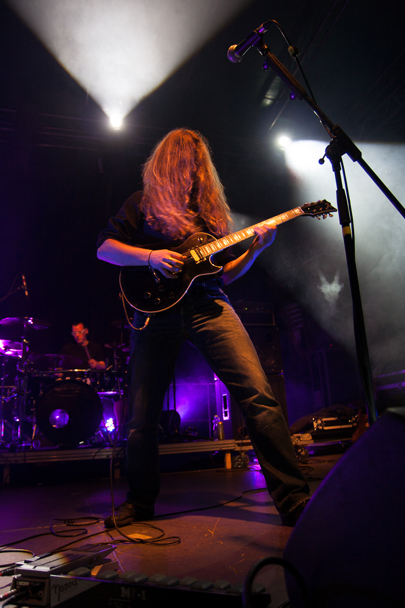 A guitarist with long hair covering their face is passionately playing an electric guitar on a dimly lit stage at the 2012 Kilkim Žaibu festival. The musician, dressed in dark clothes, stands near a microphone while a drummer can be seen in the background, both illuminated by purple and white stage lights.