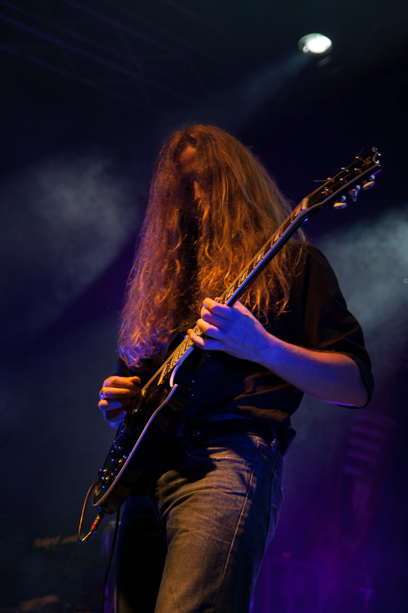 A person with long, flowing hair plays an electric guitar on stage in a dimly lit, smoky environment at Kilkim Žaibu festival. They wear a dark shirt with the sleeves rolled up and blue jeans. The scene is filled with dramatic lighting, highlighting the intense focus and dynamic presence of the musician.