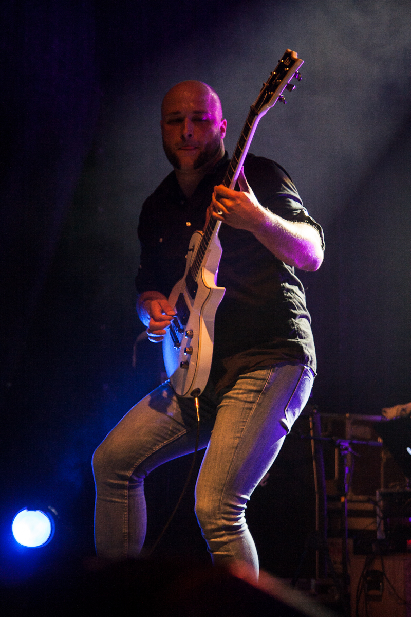 A bald person is energetically playing an electric guitar on stage at the 2012 Kilkim Žaibu festival, illuminated by dramatic concert lighting. They are wearing a dark shirt and jeans, fully engaged in the performance. A spotlight shines from the lower left, casting dynamic shadows against a dim and blurred background featuring indistinct equipment.
