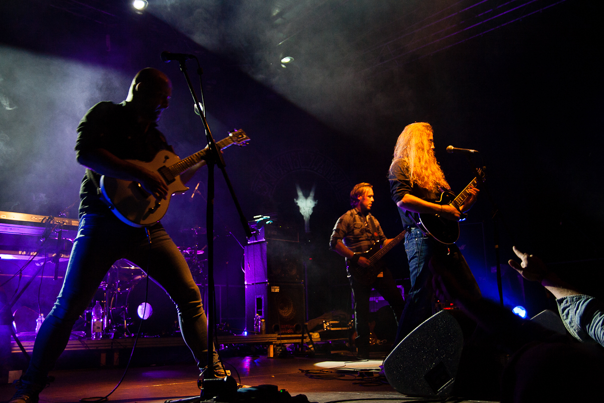 At the Kilkim Žaibu 2012 festival, a rock band performs energetically on a dimly lit stage with purple and blue lighting. The lead guitarist with long hair plays passionately while three other band members play guitar, bass, and keyboard. A faint image of a winged figure is visible on the backdrop.