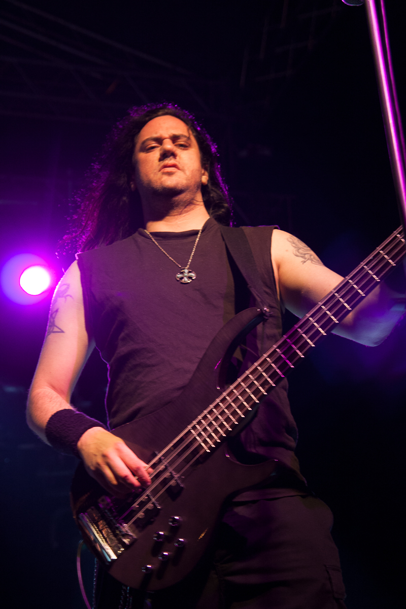 A musician with long black hair and tattoos plays an electric bass guitar on stage at the 2012 Kilkim Žaibu festival. He is illuminated by purple stage lights and wears a sleeveless black shirt, a necklace with a cross, and a wristband. The background includes stage equipment and lighting fixtures.