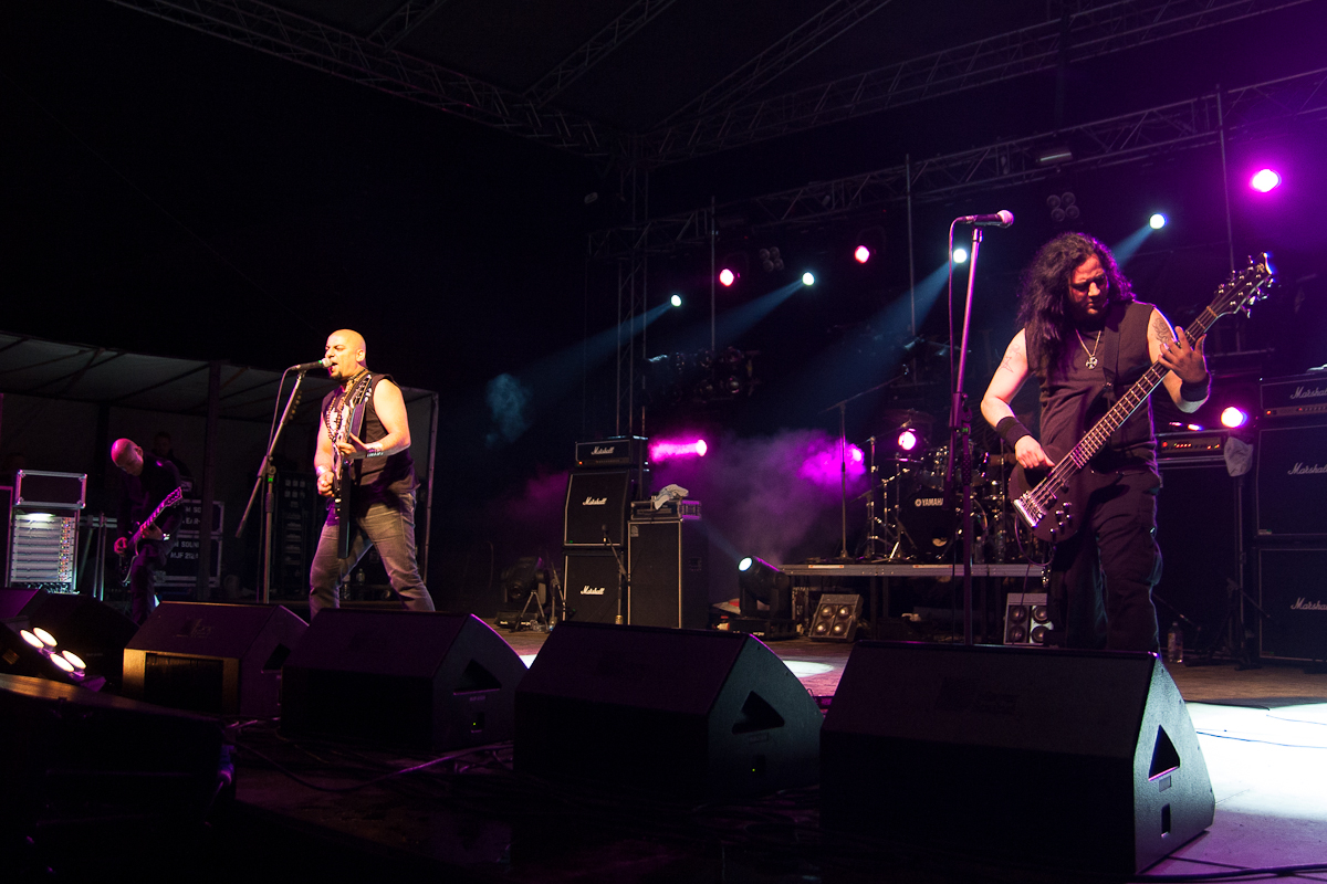 At the 2012 Kilkim Žaibu festival, a rock band performs live on stage at night, illuminated by vibrant purple and white lights. The lead singer, in a sleeveless shirt, plays guitar and sings into the mic. To his right, a long-haired bassist is engrossed in playing. Large speaker stacks and amplifiers surround them.